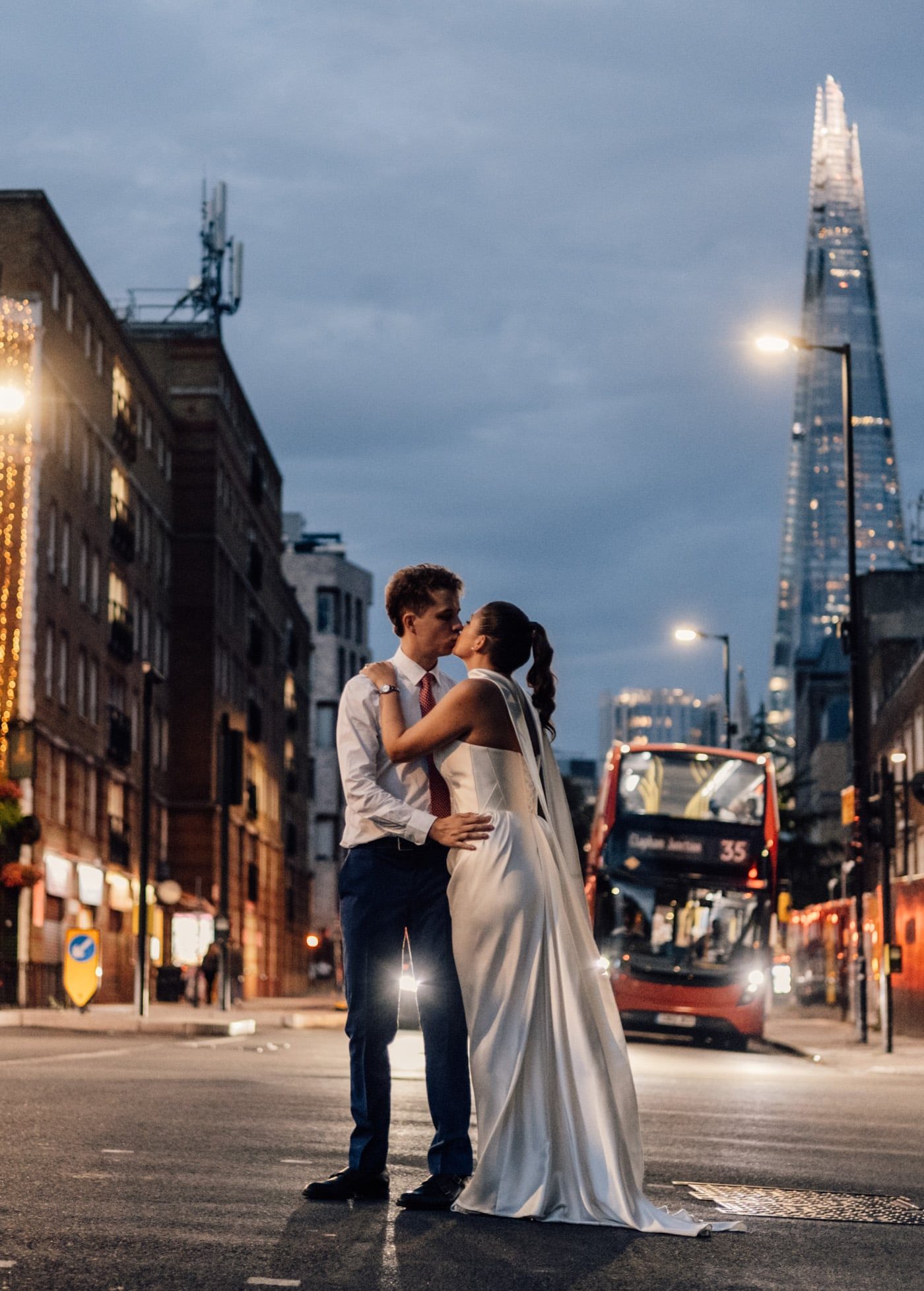 A couple kissing on a London street at dusk, with a red bus and the Shard behind them